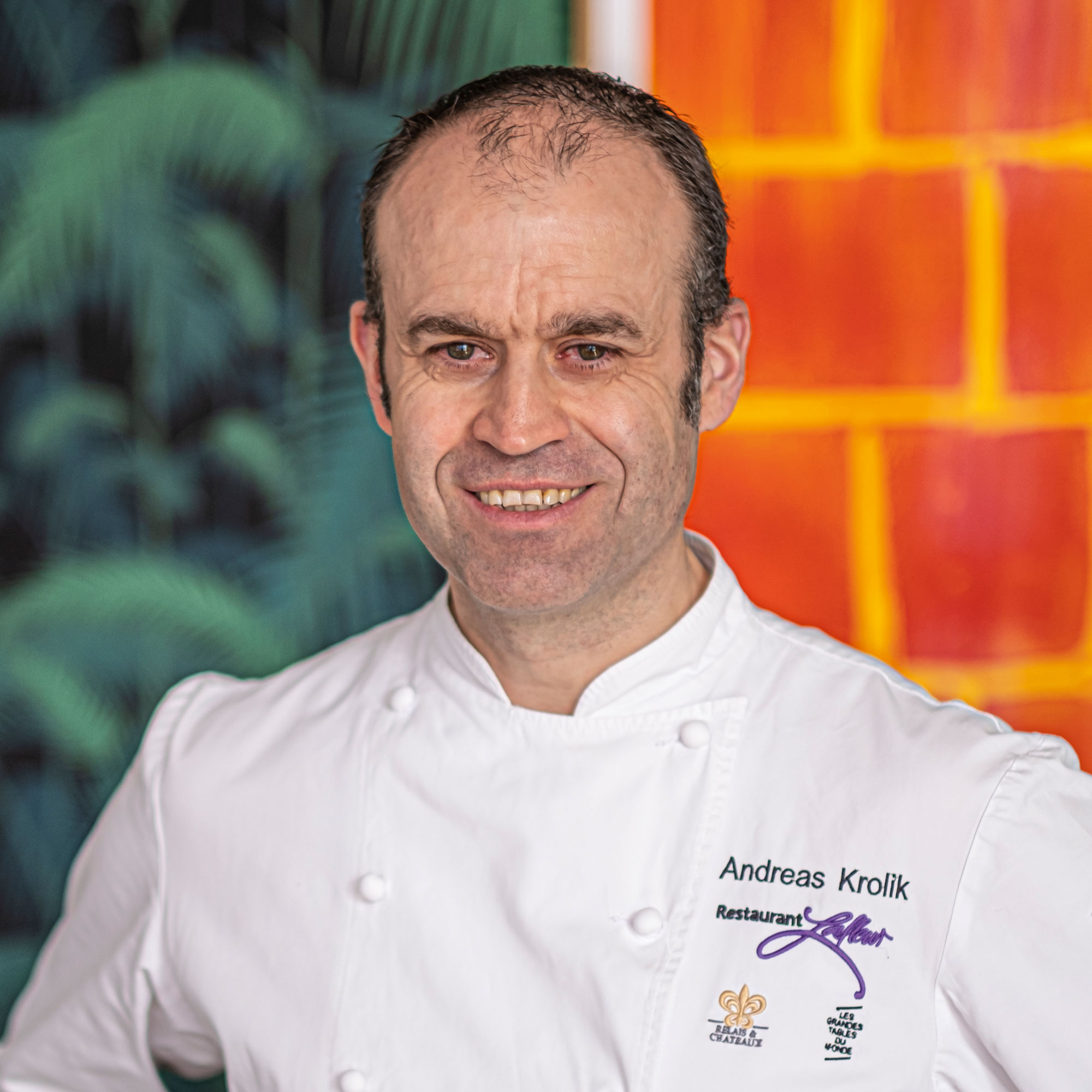Koch in weißer Jacke mit Schriftzug „Andreas Krolik Restaurant Lafleur“ vor farbigem Hintergrund