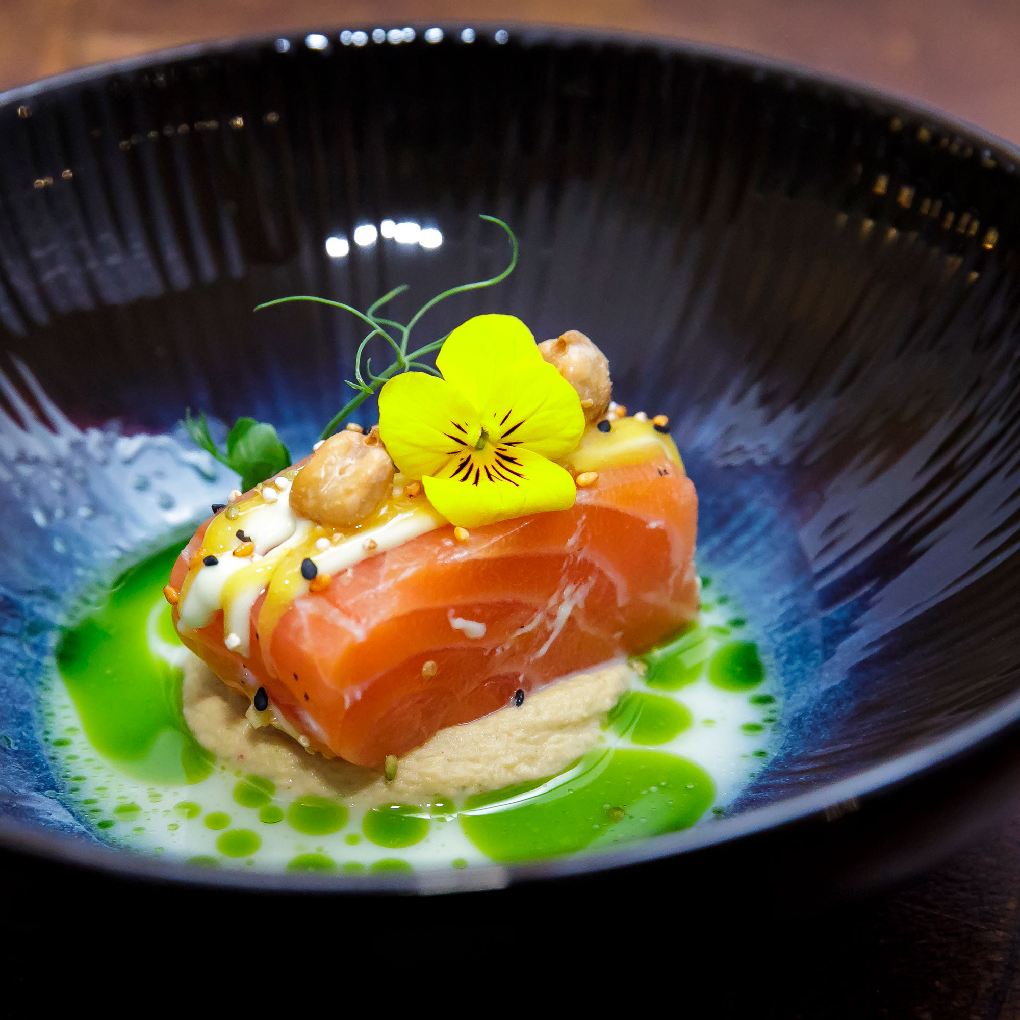 Lachsfilet auf heller Creme, garniert mit gelber Blüte, Nüssen und grüner Sauce in schwarzer Schale angerichtet