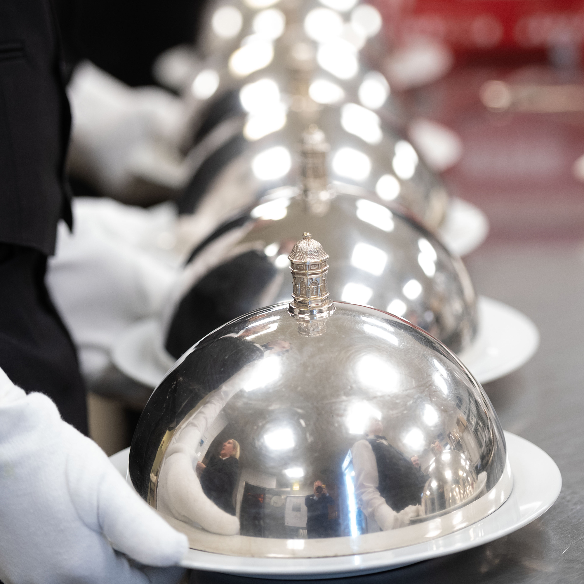 Reihe silberner Cloche-Glocken mit Schloss-Bensberg-Emblem, von Servicekräften mit weißen Handschuhen getragen
