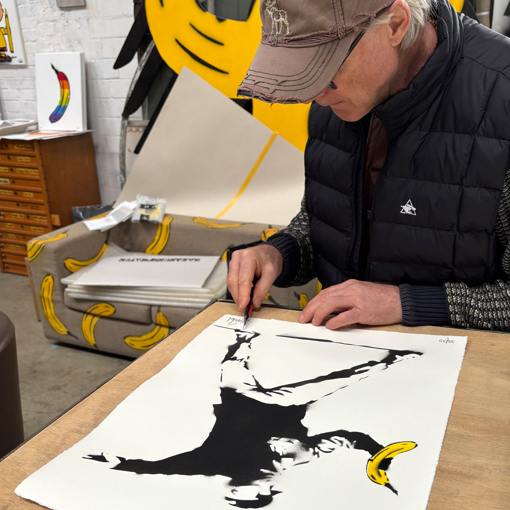 Künstler signiert Druck „Banksy-Banane“ mit schwarz-gelber Banane, im Atelier mit bananengemustertem Sessel im Hintergrund