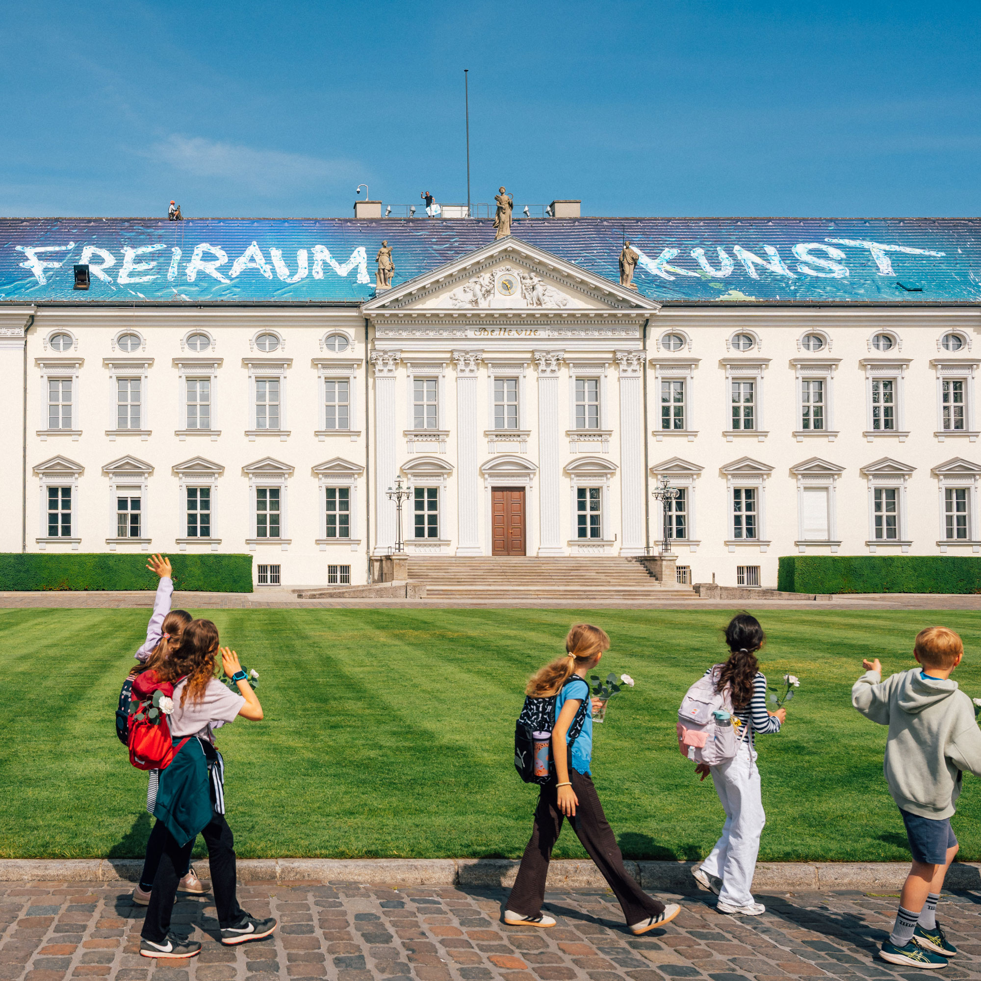 Kinder mit weißen Rosen gehen vor Schloss Bellevue, dessen Dach mit Christian Awes Kunstwerk „alive“ in Blau gestaltet ist