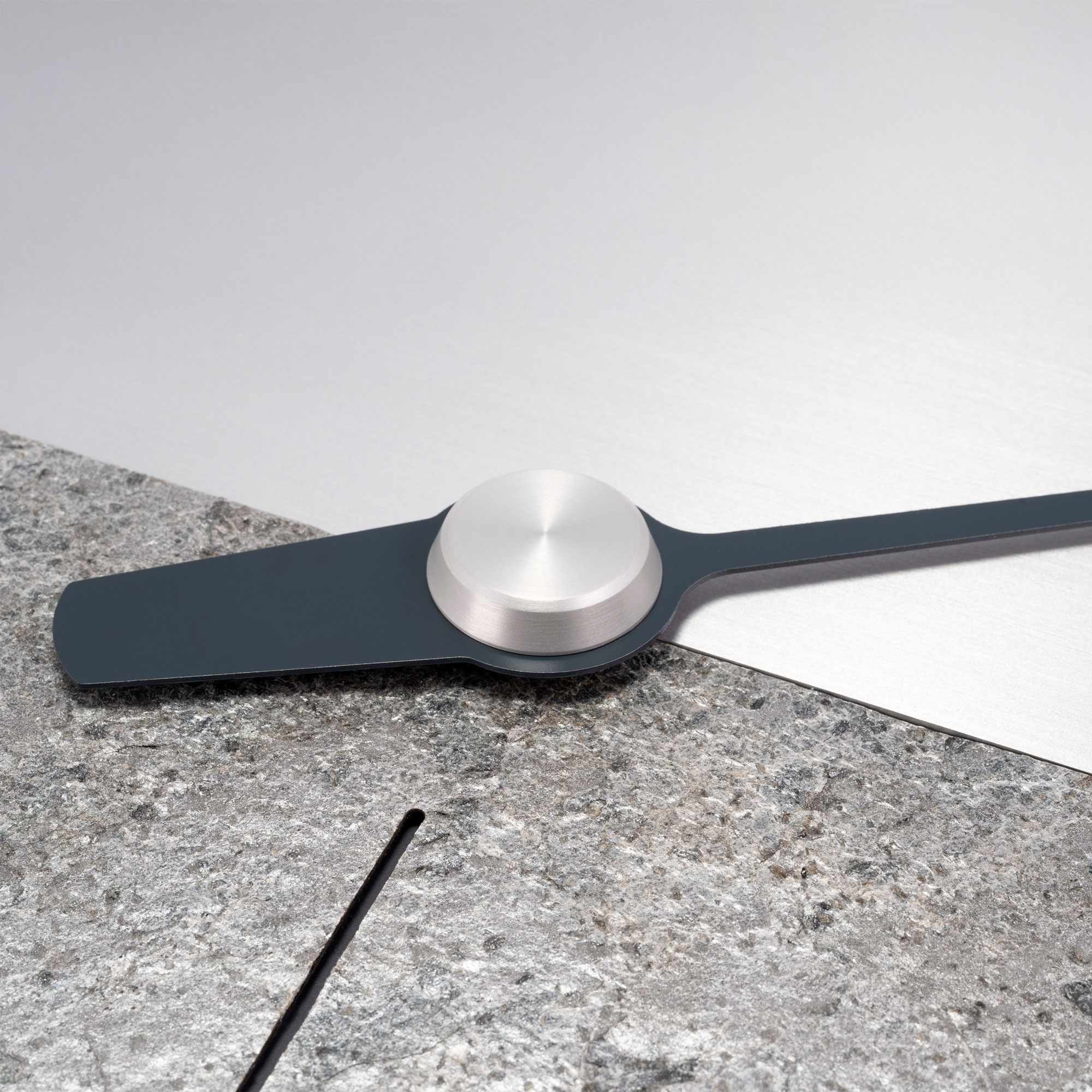 Minimalistischer Zeiger der UNO 24 Aerolith Wanduhr mit silberner Metallkappe auf strukturierter Steinfläche