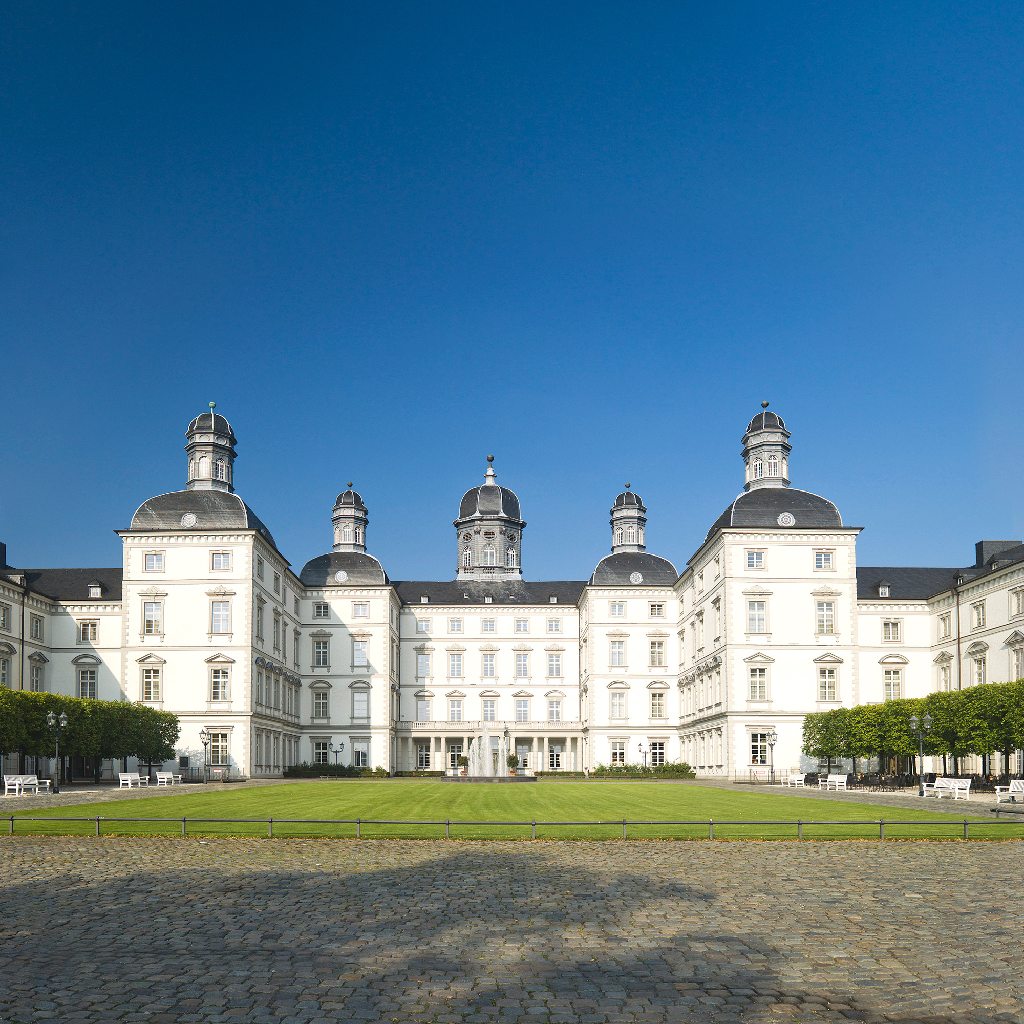 Schloss Bensberg mit weißer Fassade, vier Türmen und gepflegtem Innenhof unter klarem blauen Himmel